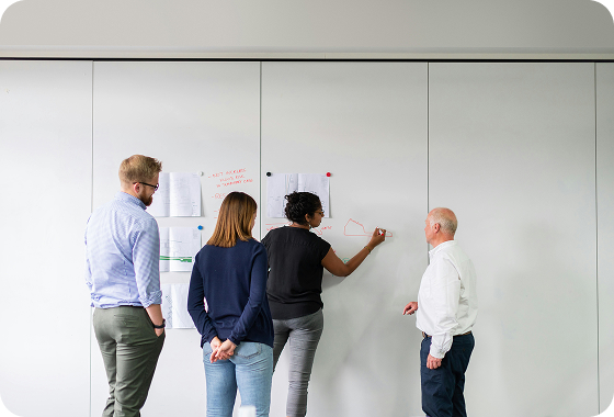 Four people stand in front of a whiteboard wall with papers and drawings pinned to it. One person is drawing on the board while the others watch and discuss.
