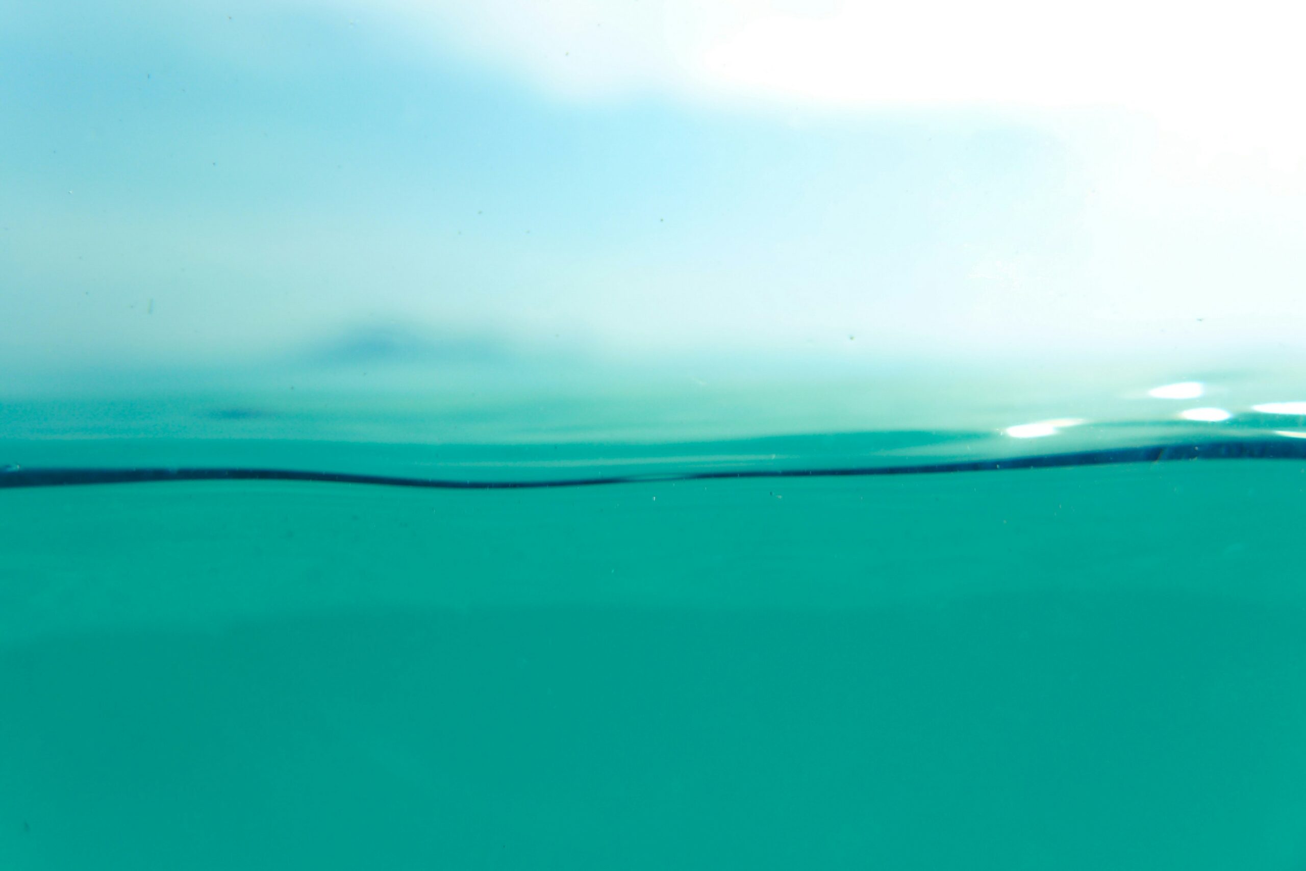 A close-up view of a calm turquoise ocean surface, with the waterline dividing the image. The sky above is bright and slightly blurred, creating a serene and tranquil atmosphere.
